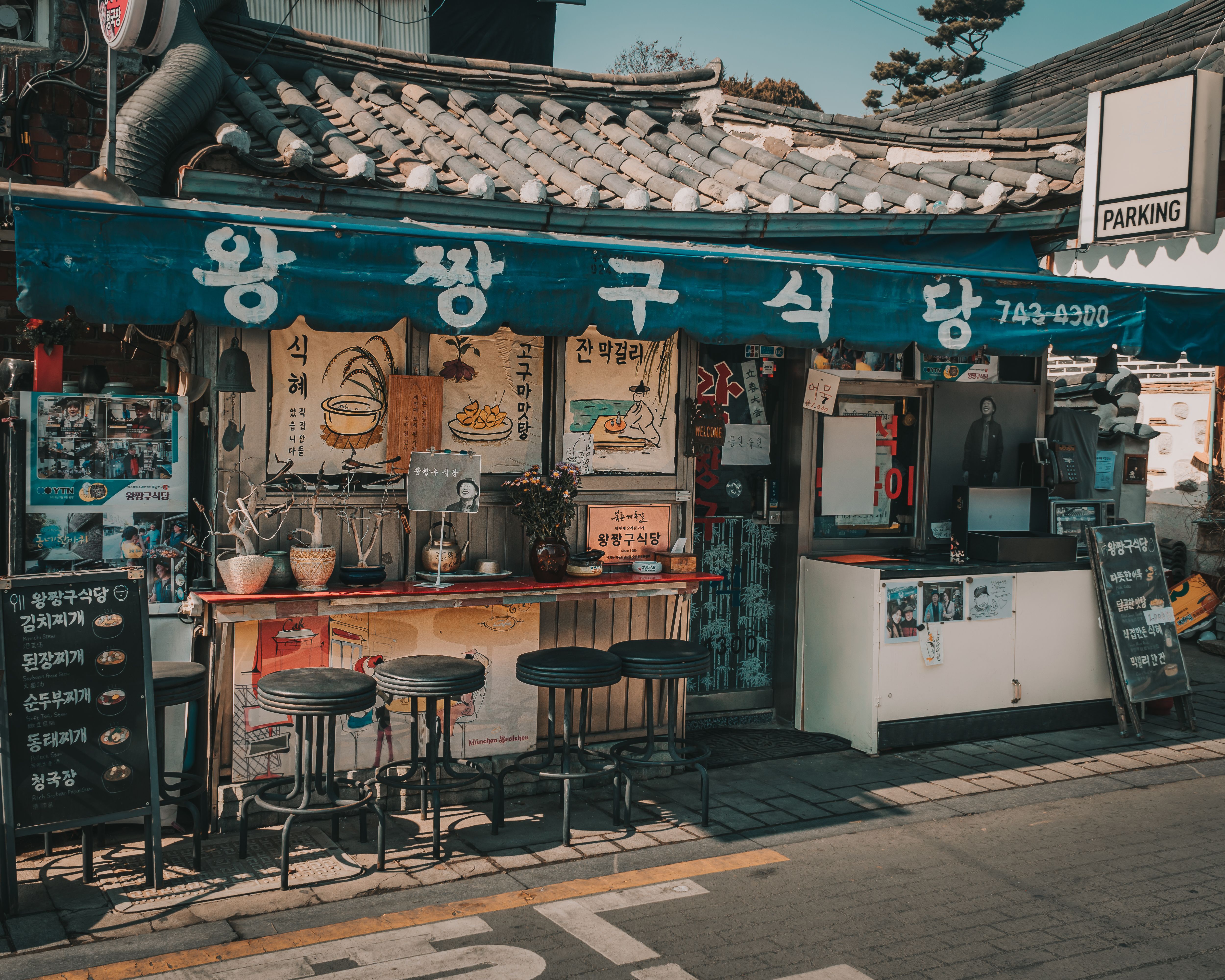 Traditional Korean restaurant Wang Pyeong Gu Sik Dang with blue awning, outdoor wooden counter, black stools, and hand-drawn menu posters in a charming street setting