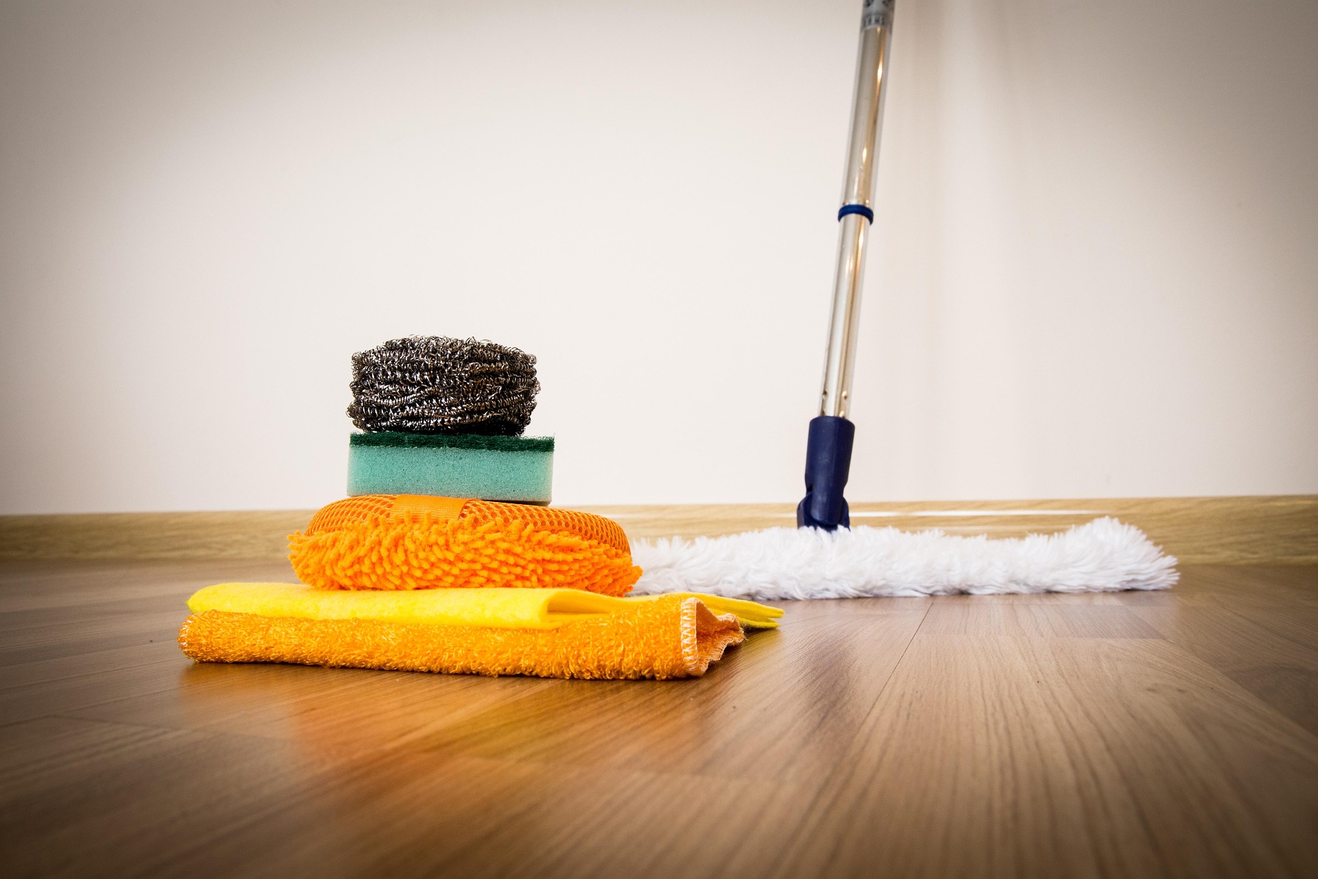 Cleaning tools, such as a sponge, a scrubber, a microfiber pad, and a mop, sit on a wooden floor, suggesting household cleaning and fresh beginnings