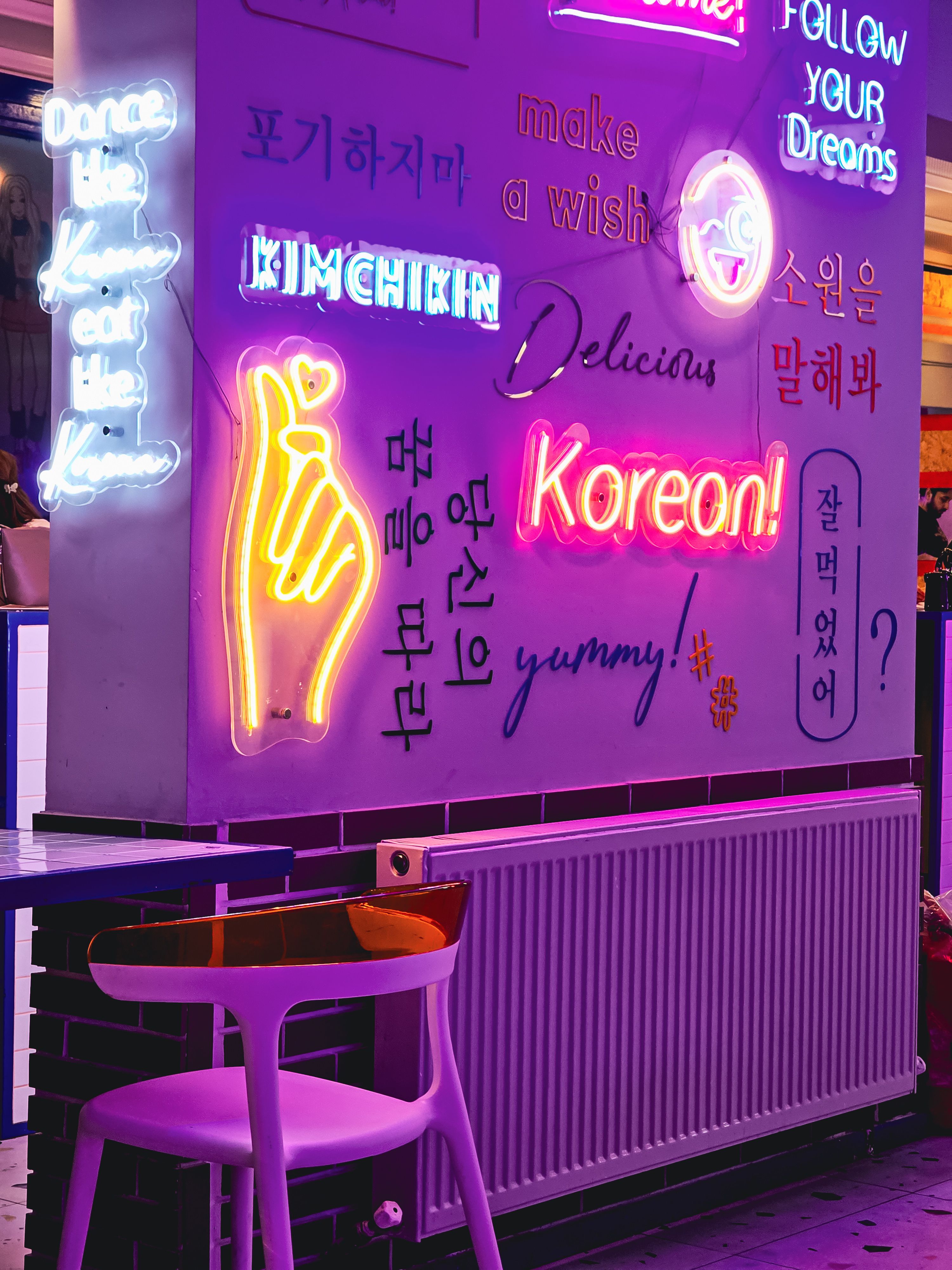 A purple neon-lit Korean-themed café wall features glowing signs, Hangul lettering, and a yellow hand-heart light, with a chair and counter below.